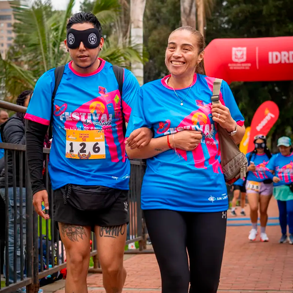 Participante con antifaz avanza tomado del brazo de una guía durante la carrera 5K Sin Límites, con otros corredores al fondo.