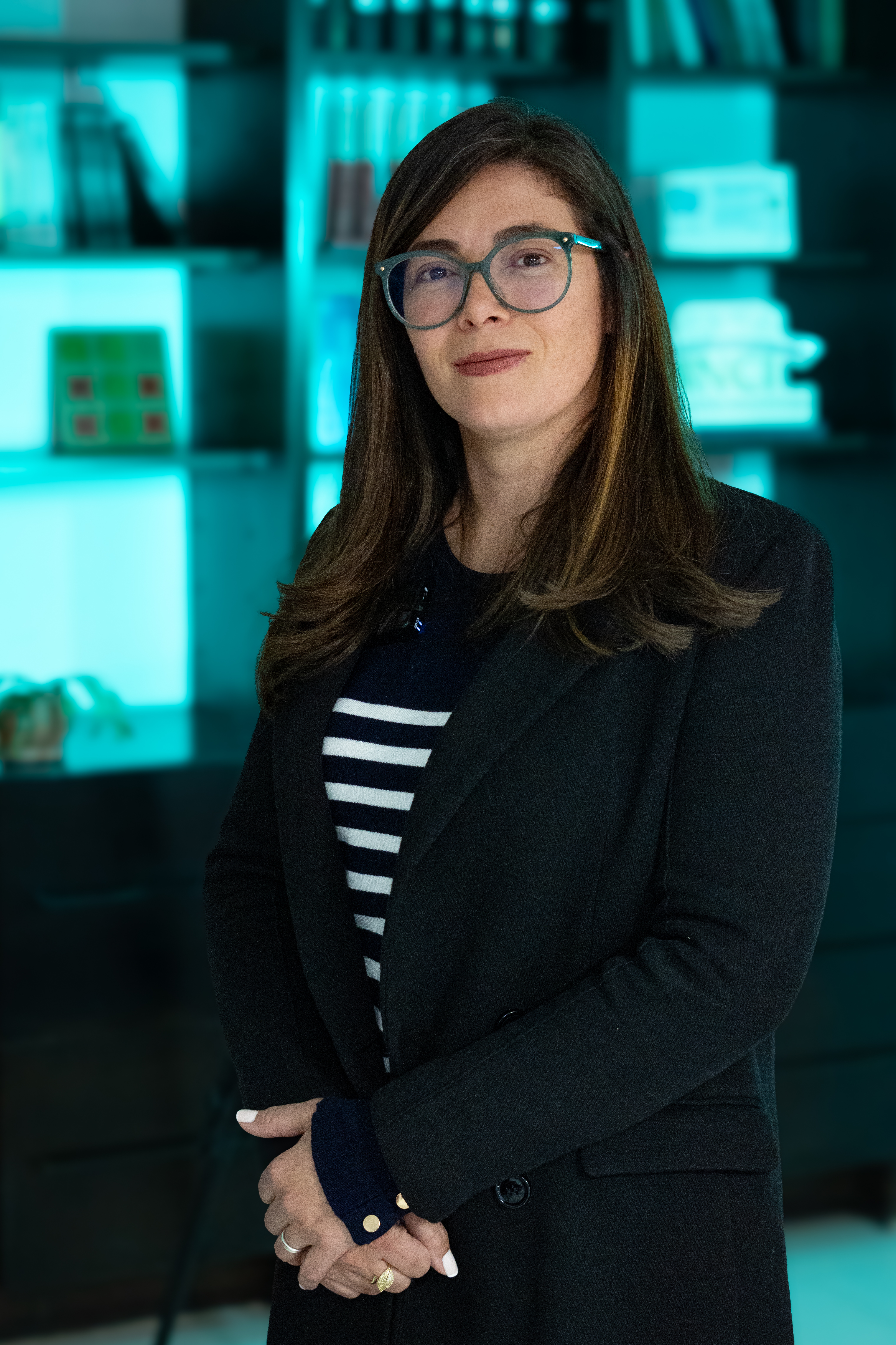 Andrea Carolina Chacón Castillo, directora general del Instituto Nacional para Ciegos (INCI). Ella está de pie en un espacio interior, frente a una estantería con libros y objetos decorativos. Tiene el cabello largo y castaño, lleva gafas de montura clara y viste un blazer oscuro sobre una camisa de rayas azul marino y blancas. El entorno está iluminado con una luz azul verdosa que aporta un aire moderno y profesional. Su postura transmite serenidad y liderazgo, en un ambiente que combina lo académico con lo institucional.