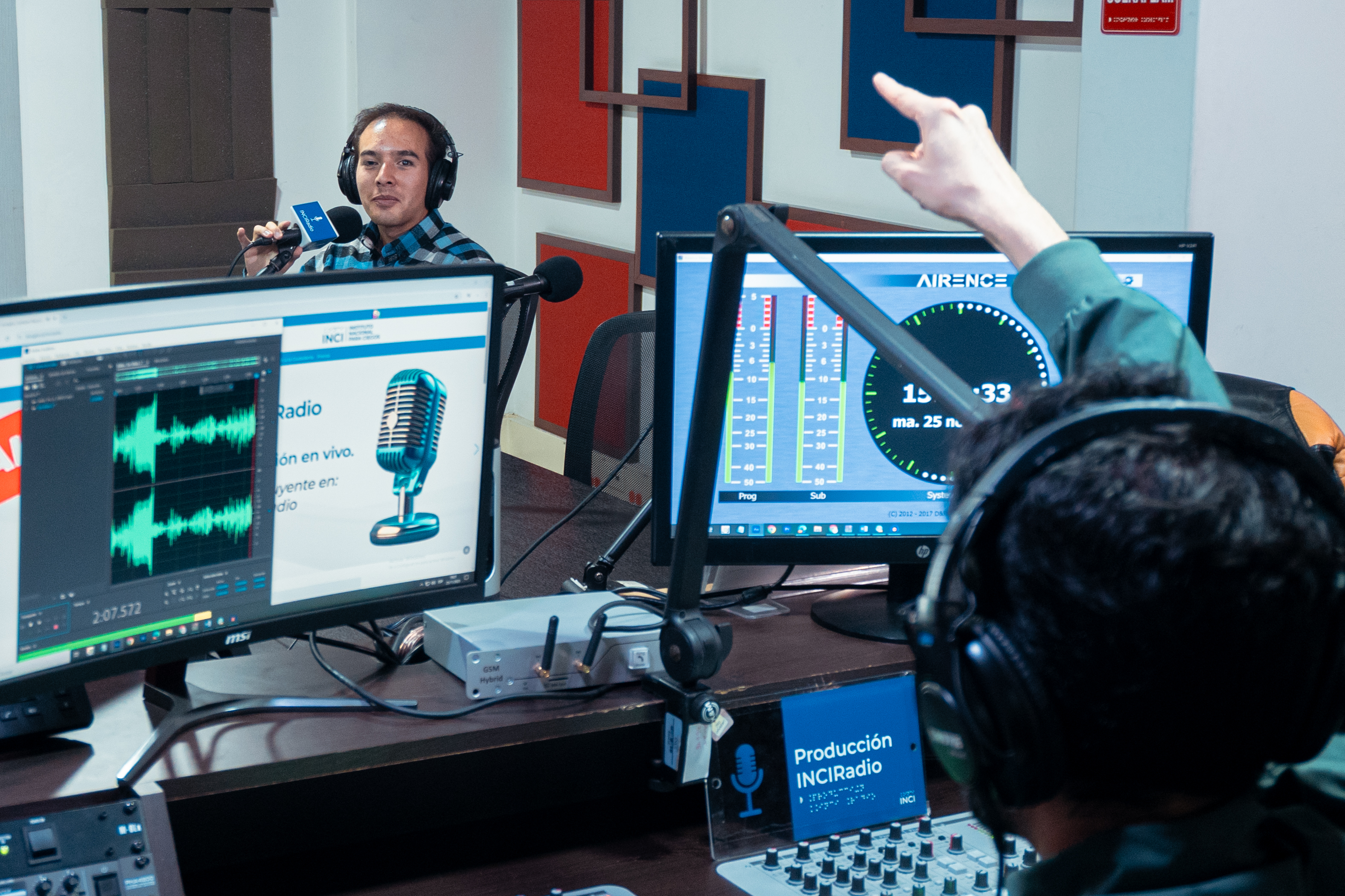 en la cabina de inciradio henry de espalda le da paso a camilo garnica quien está al fondo sentado frente al micrófono a la derecha de la foto, camilo sonríe 