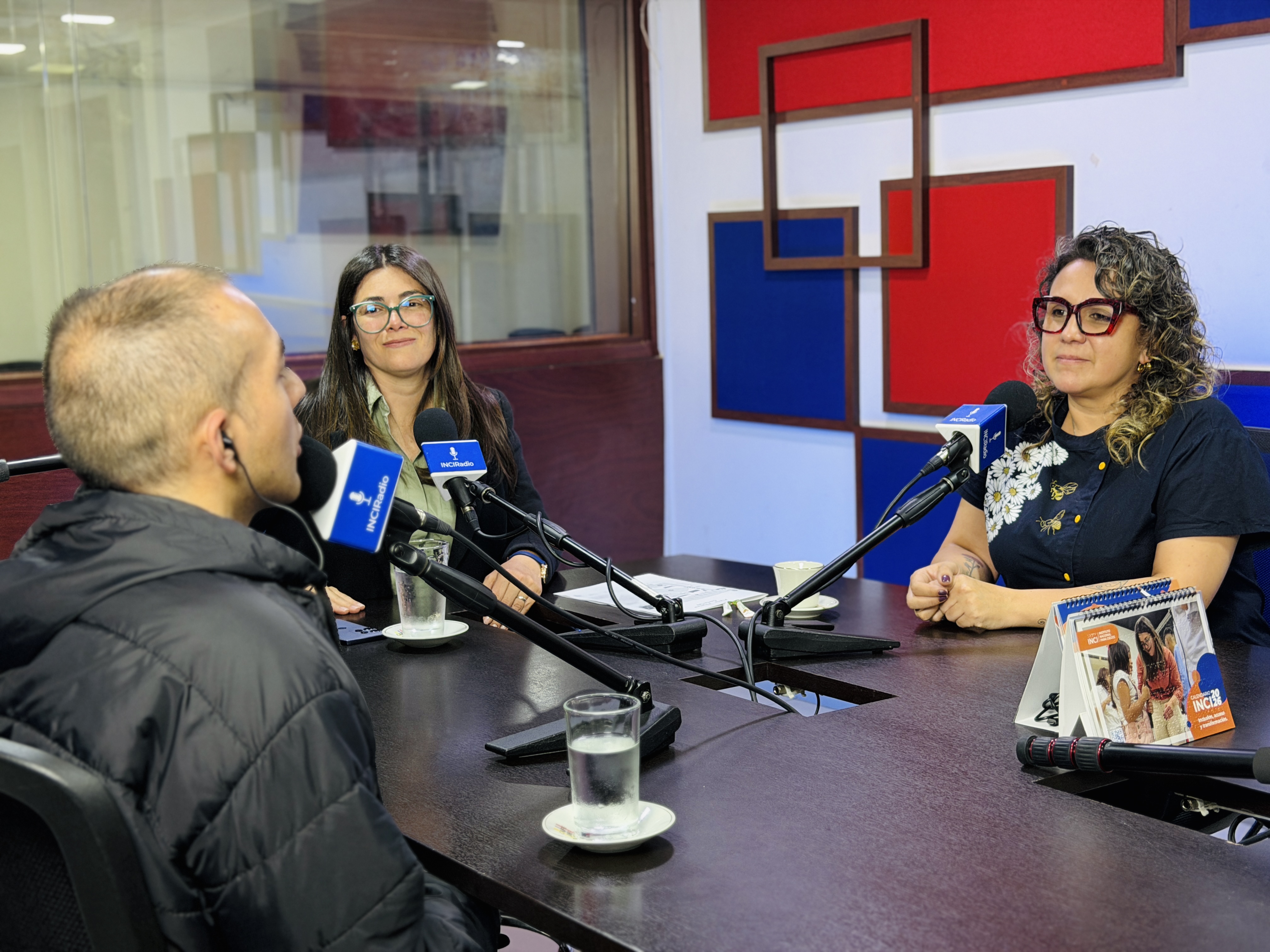 En la cabina de INCIRadio,  tres personas participan en una conversación frente a micrófonos con el logo de la emisora. A la izquierda, de espaldas, está Camilo Garnica, con audífonos puestos y un gesto atento hacia quienes tiene enfrente. En el centro, la directora general del inci  Andrea Carolina Chacón Castillo, directora general del INCI, con gafas y un blazer oscuro, A la derecha, está Adriana Ángel, directora de la Feria Internacional del Libro de Bogotá (FILBo), con gafas y una blusa oscura de diseño floral, participando activamente en el diálogo.