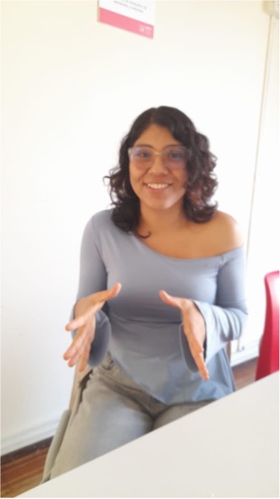 Alessandra Baltazar está sentada frente a una mesa en un espacio interior. Lleva gafas, una blusa azul de manga larga y pantalón claro. Tiene las manos levantadas al frente, realizando un gesto durante una conversación. Al fondo se observa una pared blanca y una silla roja.