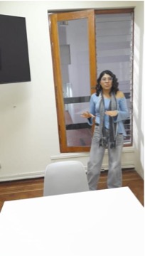 Alessandra Baltazar participa en una conversación de pie dentro de un salón. Mantiene ambos brazos extendidos hacia adelante, con las manos abiertas, acompañando sus palabras mediante gestos amplios. La imagen captura un momento de explicación e interacción, mostrando el movimiento natural de su cuerpo mientras se comunica.