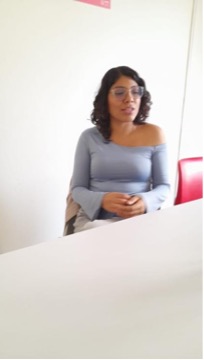 Alessandra Baltazar aparece sentada junto a una mesa, con las manos reunidas frente a su cuerpo. La imagen la muestra de perfil parcial, orientada hacia uno de los lados del espacio. A su costado se observa una silla y al fondo una pared clara que ocupa la mayor parte de la escena.