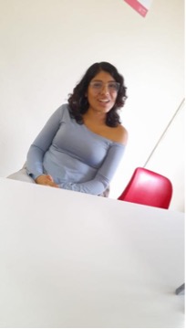 Alessandra Baltazar aparece sentada al otro lado de una mesa. Su cuerpo está ligeramente inclinado hacia un costado y dirige el rostro hacia la cámara. La imagen captura un momento de conversación, con una expresión espontánea y cercana. A su lado se observa una silla roja y, al fondo, una pared clara.