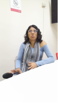 Alessandra Baltazar participa en una conversación mientras acompaña sus palabras con las manos. Una mano se extiende hacia adelante con la palma abierta y la otra descansa sobre el abdomen, reforzando la explicación que realiza en ese momento. La imagen captura el movimiento natural de sus gestos durante la interacción.