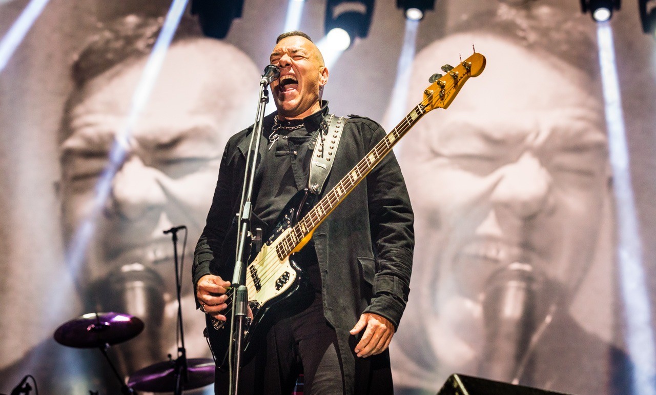 Fernando ruiz esta frente al microfono con gestos de estar cantando unas notas altas atras estan los audiovisuales del concierto que enfocan su rostro