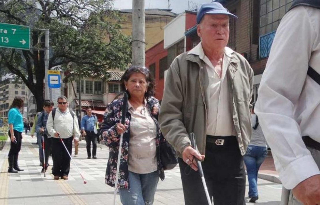 Varias personas con discapacidad visual caminando por un anden con baldosas podo táctiles