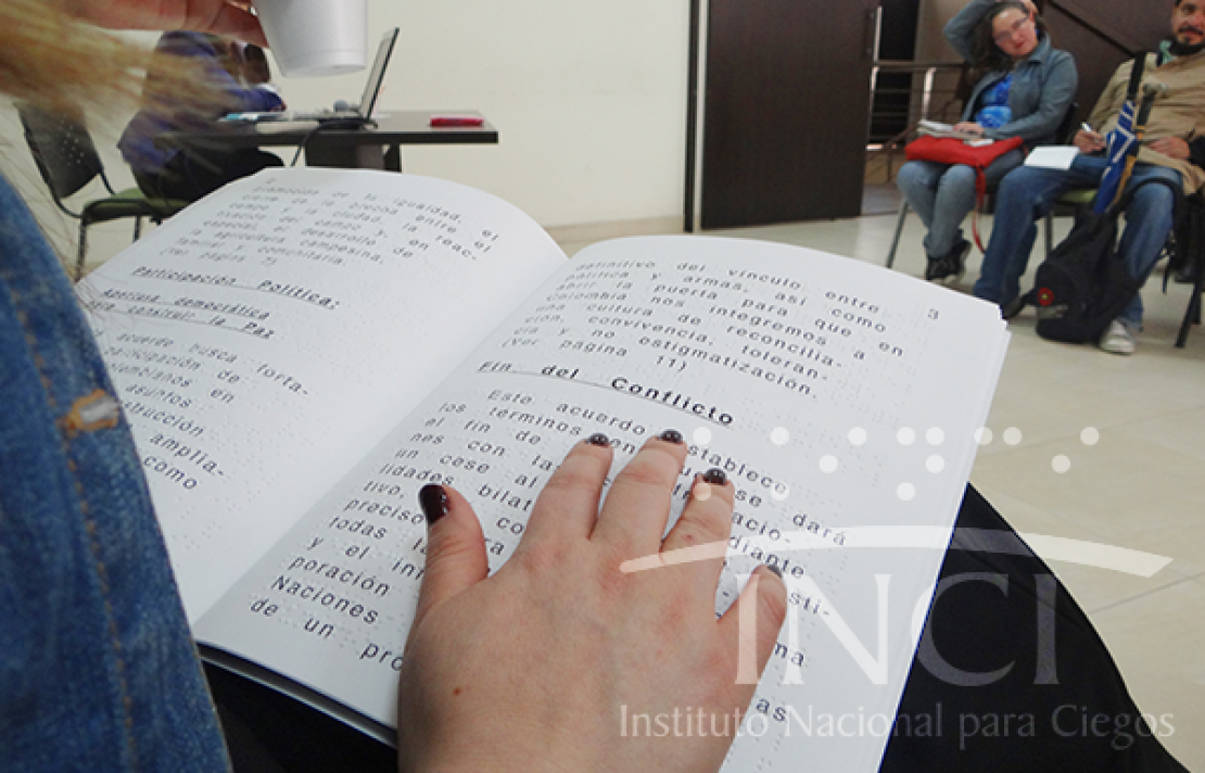una mujer sentada leyendo un texto en braille delante de un grupo de personas en un auditorio