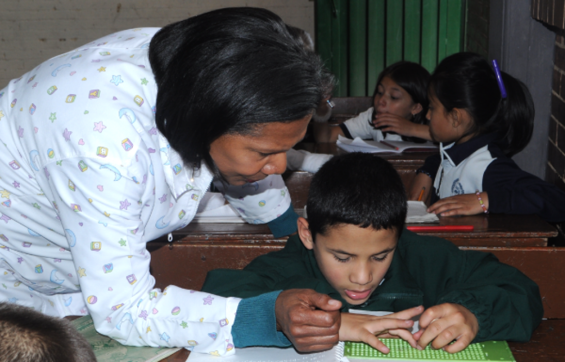 docente de apoyo enseñando a un niño con discapacidad visual a utilizar una pizarra plástica sencilla