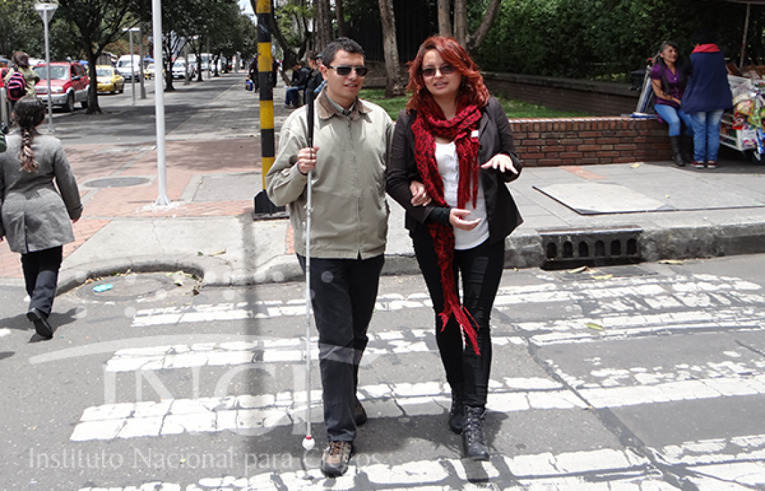 un joven con discapacidad visual cruzando una cebra en compañía de su bastón y una persona guía y su