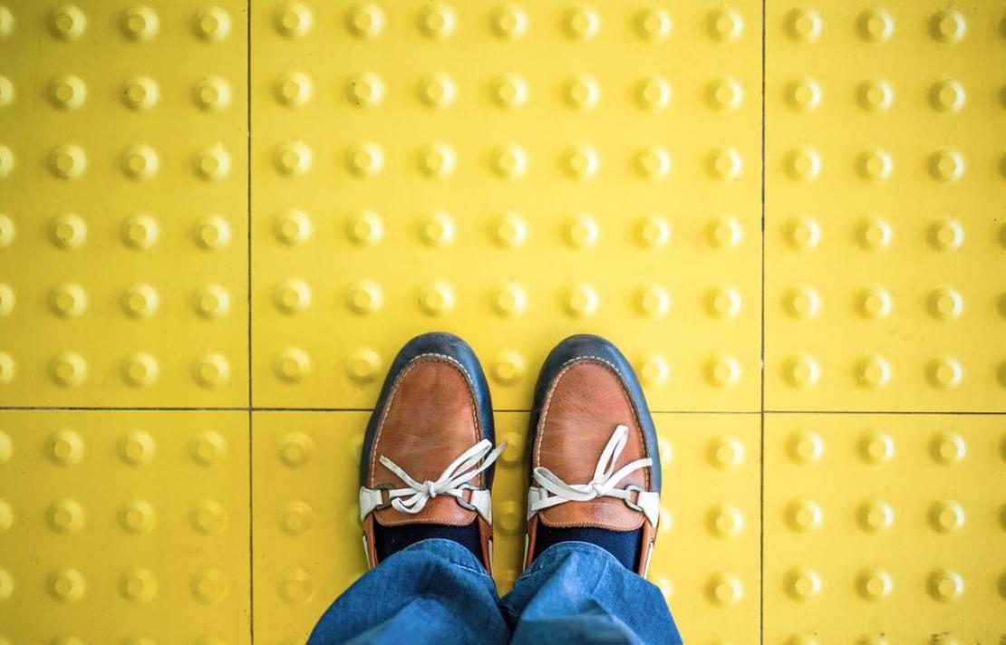 señal podotactil imagen de una señal podotáctile, sobre ella aparecen las piernas de una persona que viste un jean de color azul, sus zapatos son café