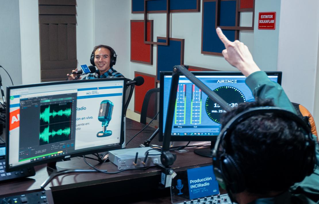 camilo garnica está frente al microfono, mientras un hombre con chamarra verde le hace la señal que va al aire, Camilo sonríe