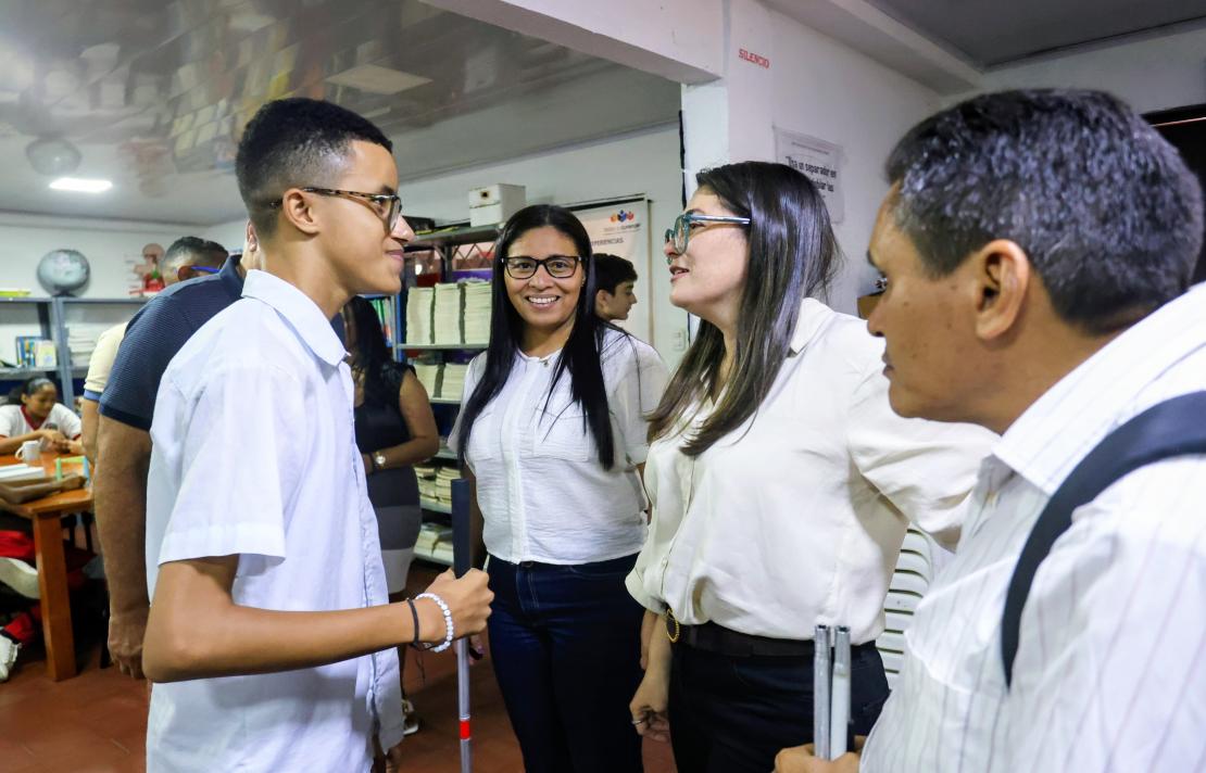 En un aula, la directora del INCI, conversa de cerca con un estudiante con discapacidad visual, quién sostiene un bastón, mientras otras personas observan alrededor.
