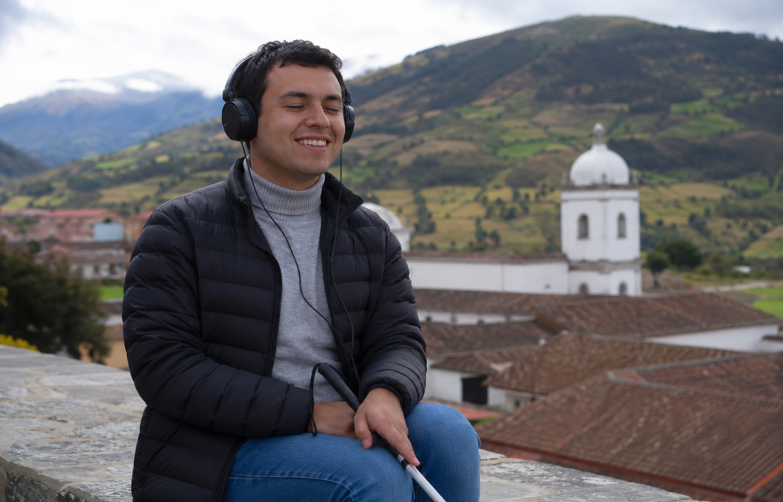 en Boyacá en la plaza una persona ciega sonríe usa audífonos en sus manos tiene un bastón blanco