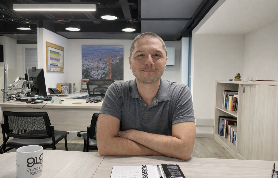 en la oficina de comunicaciones de javeriana estereo Esteban Bernal Carrasquilla sonríe, el tiene el pelo corto, está cruzado de brazos y viste una camisa de color gris