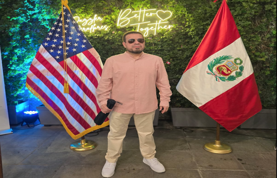 Braulio viste una camisa rosada un pantalón beige, tenis blancos, detrás de él está la bandera de estados unidos y perú