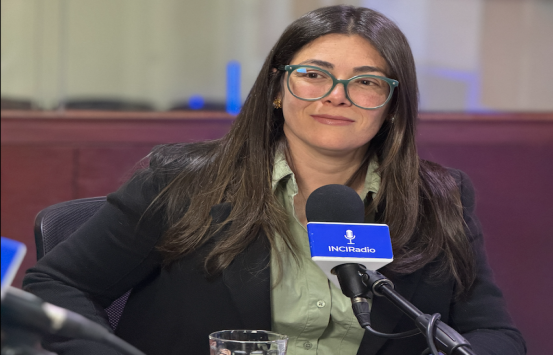 Andrea Carolina Chacón Castillo, directora general del Instituto Nacional para Ciegos (INCI),  está sentada frente al  escritorio de transmisión, hablando en un micrófono que lleva el logo de la emisora. Viste una camisa verde claro y un blazer oscuro, y usa gafas de aumento.