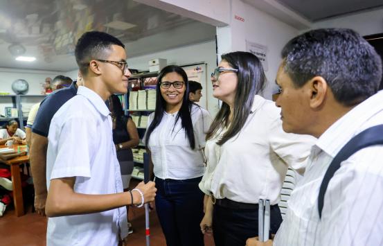 En un aula, la directora del INCI, conversa de cerca con un estudiante con discapacidad visual, quién sostiene un bastón, mientras otras personas observan alrededor.