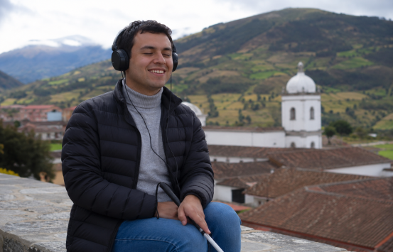 en Boyacá en la plaza una persona ciega sonríe usa audífonos en sus manos tiene un bastón blanco