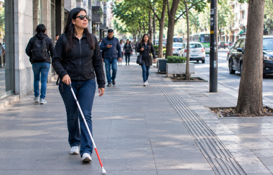 Mujer con discapacidad visual se desplaza de manera autónoma por una acera de la ciudad utilizando bastón blanco.