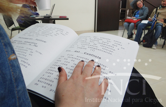 una mujer sentada leyendo un texto en braille delante de un grupo de personas en un auditorio