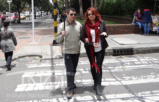 un joven con discapacidad visual cruzando una cebra en compañía de su bastón y una persona guía y su